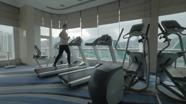 Steadicam shot of young woman running on treadmill in modern gym with panoramic windows. Sport training and enjoying cityscape from upper floor