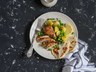 Lemon thyme baked chicken, potatoes and green peas on a white plate on a dark background. Delicious lunch