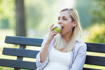Fototapeta premium Young happy woman is sitting in park and eating apple.