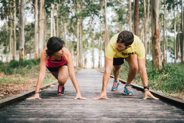 Fitness couple ready for running race on wooden walkway on an outdoor park looking each other challenging in powerful confident starting line pose in Rodiles, Asturias.