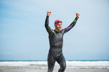 Fit sporty open water swimmer man running off shore on a beach rising arms up in victory sign after swimming triathlon competition exercise routine workout. - obrazy, fototapety, plakaty