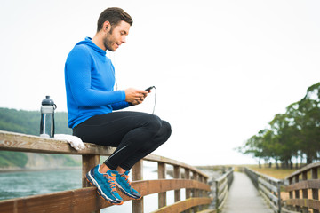 Outdoor runner man sitting on wooden walkway listening looking and touching his phone with water bottle and white towel. Fitness fit male motivated for outside workout exercises.