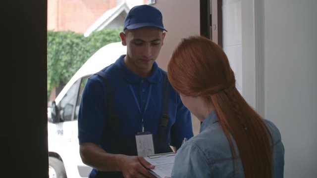 Delivery man entering house of young woman customer, she signing documents and receiving a parcel