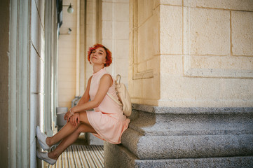 woman with dyed red hair in a pale pink dress with white backpack, sitting between columns holding up the legs