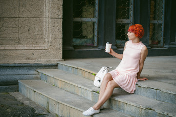 He drinks coffee from a cup. woman with dyed red hair in a pale pink dress drinking coffee, holding a paper cup of coffee to go, sitting on the steps outside a summer day.