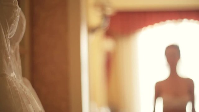 Beautiful white wedding dress is hanging on a hanger in a bedroom , amazing bride with a crown and luxury jewel le ry comes to the dress in a bright sunshine and tries it on, she looks satisfied.