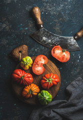 Fresh colorful ripe heirloom tomatoes on wooden board, herb chopper knife for cooking over grunge dark plywood background, top view. Harvest vegetable cooking conception.