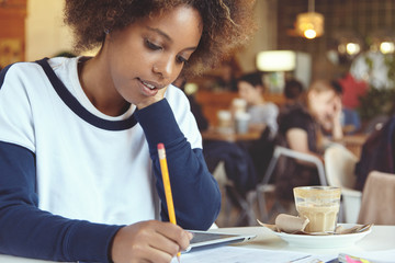 Beautiful African graduate student girl holding pencil, writing down in sheet of paper while preparing for final exams at university, using touch pad, resting her elbow on table at cafeteria