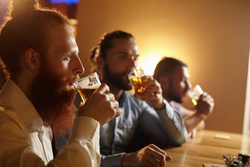 Three handsome bearded Caucasian men holding glasses, drinking light beer, sitting at bar counter and watching football. Colleagues relaxing at pub after hard working day, enjoying cold alcohol drinks