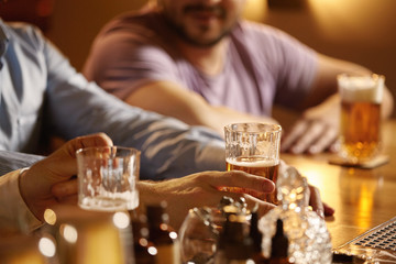 Happy young businessmen drinking beer and talking at pub after work. Cropped shot of three young men in casual wear toasting with beer while sitting at table, holding glasses of lager or ale