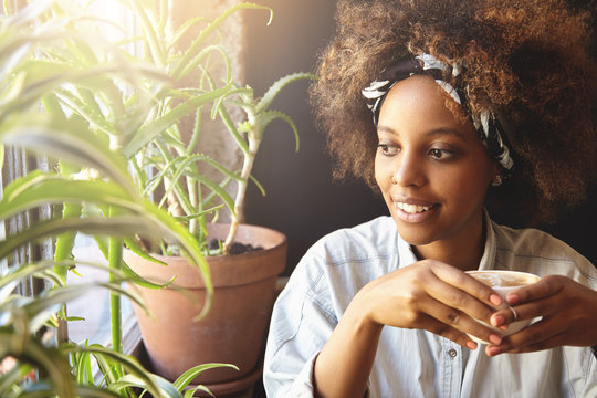 Portrait of young hipster dark-skinned girl with nose-ring and do-rag on her head, enjoying hot coffee or tea, looking through window with pensive expression, thinking and making plans for day