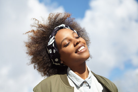 Close up shot of young black hipster student wearing do-rag on her head, smiling and closing her eyes with happy face expression, dreaming and enjoying warm summer wind while walking outdoors alone
