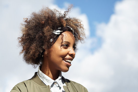 Headshot of beautiful dark-skinned young woman with Afro haircut wearing bandana and stylish clothes, relaxing outdoors, posing against blue sky background with copy space for your advertising content