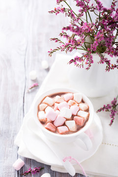 Cup of hot chocolate with mini marshmallows with pink flowers in jug on white wooden background with copy space.