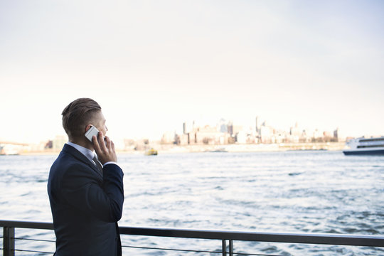 Businessman talking on smart phone while standing by river against clear sky