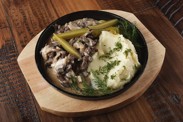 Beef stroganov with mushed potatoes and pickled cucumbers in a pan. Wooden background.