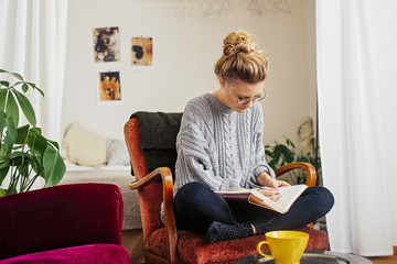 Woman writing on book while sitting on chair at home