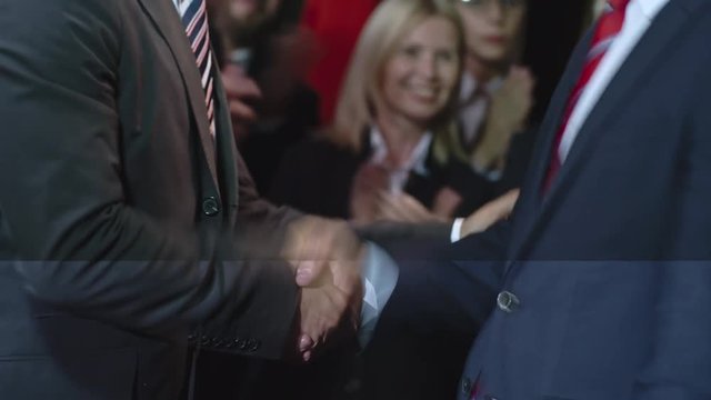 Mid-section of two politicians meeting at political convention and shaking hands as pleased audience supports them with applause 