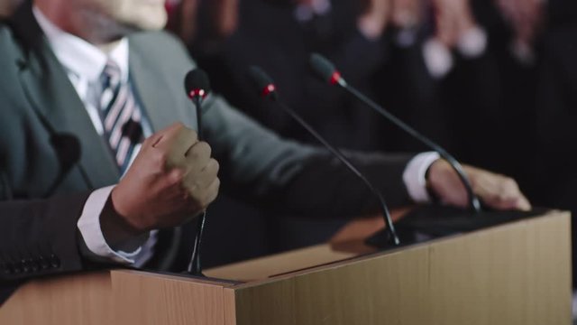 Closeup with selective focus of male politician hands lying on tribune as he delivering passionate speech before public, then making a fist and wildly gesturing