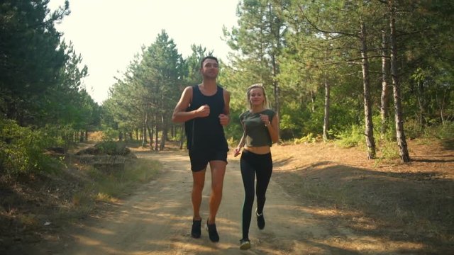 Sportive couple in sportswear smiling jogging along path in forest Slow motion