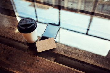 One Blank Kraft Paper Business Card Mockup Wood Table.Take Away Coffee Cup Coworking Place.Modern Phone Work Office Reflection Glass Background.Mock Up Objects Ready Private Message.Horizontal Photo.