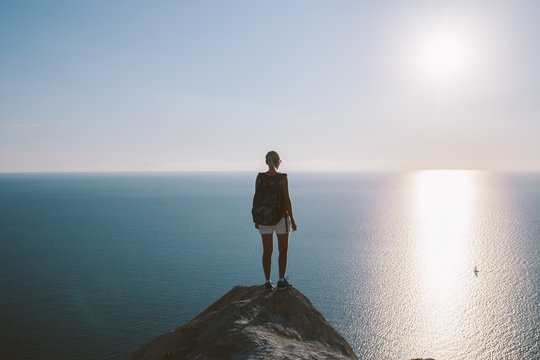 Young woman hiker with backpack standing on cliff and looking forward on the background of the sea, sky. lady tourist on top of a mountain enjoying view... - Powered by Adobe