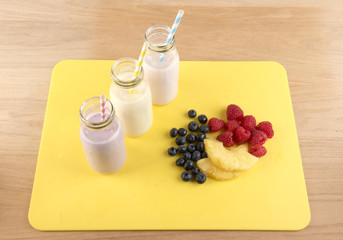 Image of three smoothies in glass bottles on a yellow chopping board, with a pile of mixed fruit. 