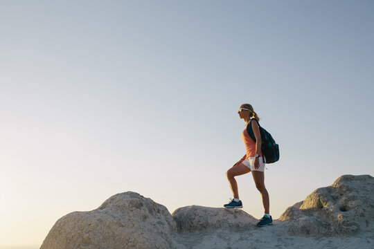Young woman hiker with backpack standing on cliff and looking forward. lady tourist on top of a mountain enjoying view before sunset. Sport lifestyle travel concept