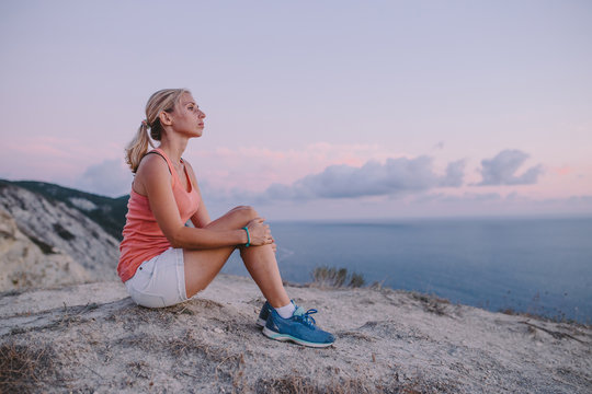 active young woman tourist sitting on top of a mountain and looking forward on the background of the sea, sky. Sport lifestyle travel concept