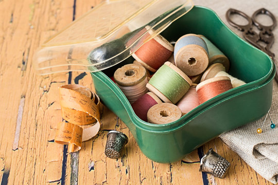 Sewing supplies,.   Box with a spools of thread, a piece of fabric, scissors and thimbles on an old wooden table.