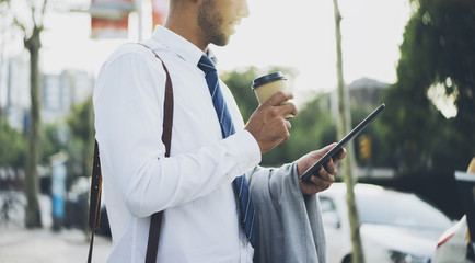 Cropped image of professional businessman using digital tablet and holding coffee to go while working at office, young manager drinking coffee at break outside with tablet pc