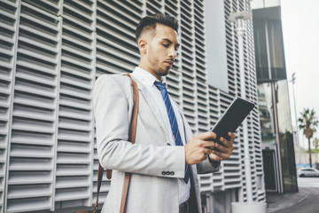 Professional young businessman wearing grey suit and using digital tablet during break, latin male lawyer searching information at internet via tablet pc, office in the background