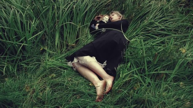 Stylized shot creepy white faced woman with a doll in the grass in the woods laying