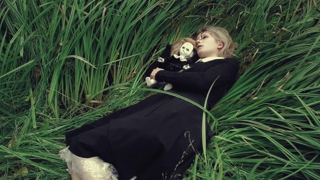 Stylized shot creepy white faced woman with a doll in the grass in the woods laying