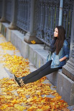 Young long-haired brunette sitting on a concrete curb around the cast-iron fence, under the feet of yellow autumn maple leaves, side view.