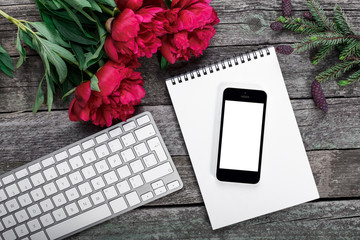Workspace with smartphone, keyboard, notepad, fir branch and peonies flowers bouquet on rustic background. Top view, flat lay