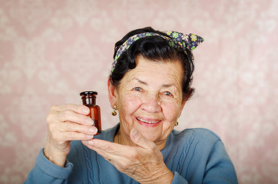 Older Cool Hispanic Woman Wearing Blue Sweater, Flower Pattern Bow On Head Holding Up A Small Red Glass Bottle And Smiling To Camera