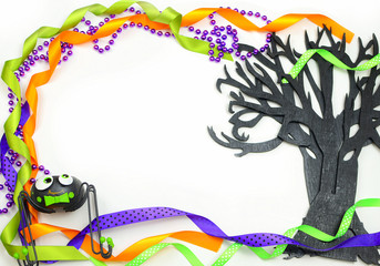 Halloween border of a rough textured wooden cutout of bare tree shape painted black. A silly spider and purple, green and orange ribbons and beads frame the copy space on white background