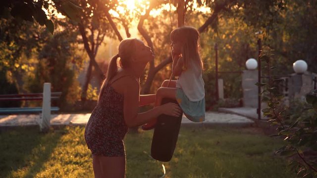 Mother tender kissing her small daughter and ride on a swing on backyard at home, in foreground and sunlight coming through trees.