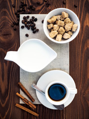 Cup of coffee, milk jug, bowl with cane sugar, cinnamon sticks and coffee beans on the gray napkin on the wooden table. Top view