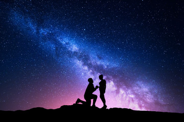 Milky Way. Night landscape with silhouettes of a man making marriage proposal to his girlfriend and starry sky. Silhouette of lovers. Couple, relationship. Milky way with people. Universe - obrazy, fototapety, plakaty