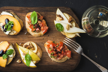 Bruschetta set with various toppings on wooden serving board and glass of white wine over black plywood background, top view, horizontal composition