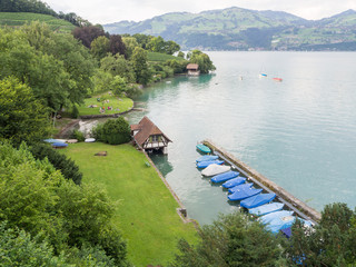 Fototapeta premium Paisaje acuático a orillas del lago Thunersee con barcas azules y casas en la orilla, rodeada de naturaleza verde y montañas al fondo, Interlaken, Suiza viajando en verano de 2016 