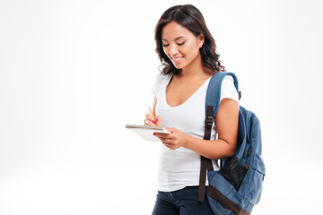 Cheerful attractive asian girl with backpack making notes in notebook