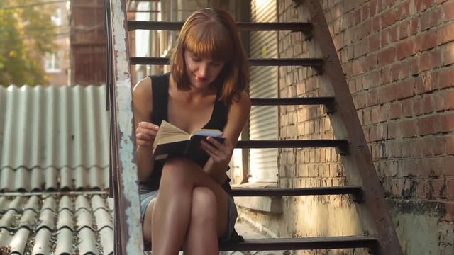 Cute girl in mini skirt reads a interesting book while sitting on the stairs at the street in a sunny evening