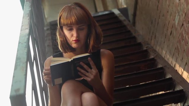 Beautiful girl in mini skirt reads a interesting book while sitting on the stairs at the street in a sunny evening