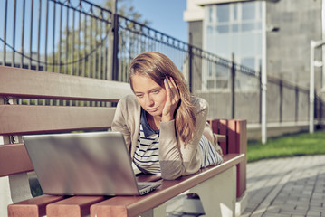 Fototapeta premium beautiful young brunette woman in a dress with blue and white stripes in sunglasses sitting on a bench and using laptop for surfing, chat, learn, game. Concept of urban social networking and internet.