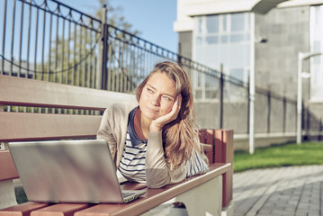 Fototapeta premium beautiful young brunette woman in a dress with blue and white stripes in sunglasses sitting on a bench and using laptop for surfing, chat, learn, game. Concept of urban social networking and internet.