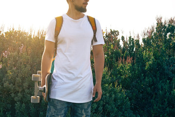 A male with a skateboard in a blank white t-shirt is standing in a park. A man with a backpack is looking aside on a plant green background.