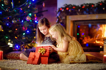 Fototapeta na wymiar Happy little sisters opening magical Christmas gift by a fireplace